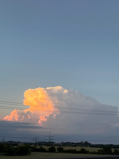 This vertical, high-angle photograph captures a dramatic sky over Wustermark, Germany, at what appears to be sunset. A large, cumulonimbus cloud formation dominates the middle ground, with its upper sections illuminated by a vibrant orange glow, suggesting it is catching the last rays of sunlight. The lower portions of the cloud are a softer pink and gray, transitioning to darker, more ominous storm clouds near the horizon.

Stretching horizontally across the image, just below the illuminated cloud, are several power lines, supported by unseen structures. In the background, silhouetted against the dusky sky, are multiple tall, lattice-like electricity pylons, characteristic of the German landscape. The foreground consists of a dark, silhouetted band of trees and foliage, with hints of agricultural fields and possibly some distant structures. The sky above the cloud is a clear, pale blue, fading to a slightly deeper hue at the top of the frame. No people or animals are visible in the image. The overall impression is one of nature's powerful display against a backdrop of human infrastructure.