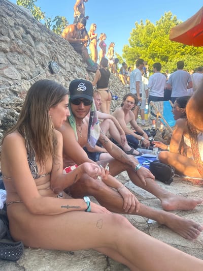 A daytime outdoor scene captures a densely populated gathering on a rocky coastal area in Tisno, Croatia. The foreground features a fair-skinned female with light brown hair, wearing a snakeskin-patterned bikini, seated facing left, conversing with a tanned male seated to her right. She has a sword tattoo on her left forearm and a cloud tattoo on her right upper thigh, and a light blue wristband on her left wrist. The male wears a black baseball cap with a white 'S' logo, sunglasses, red and white checkered swim trunks, and a light green and multicolored bandana draped over his neck. He has multiple arm and leg tattoos, several rings on his fingers, a white wristband on his left wrist, and holds a lit cigarette or joint in his right hand.

The midground and background show numerous individuals, predominantly young adults, scattered across a natural rocky slope on the left and a flatter area to the right. Many are in swimwear or casual attire. Noteworthy individuals include a dark-skinned male with dreadlocks and green socks seated on the upper rocky slope, looking at a device; a female in a black thong and black top facing away on the lower slope; and various other people standing, sitting, and interacting. A blue and white striped deck chair and a partially visible reddish-orange sun umbrella are present on the right. The environment consists of rough rock formations, green trees in the background, and a clear blue sky overhead. Natural light suggests midday or early afternoon conditions.