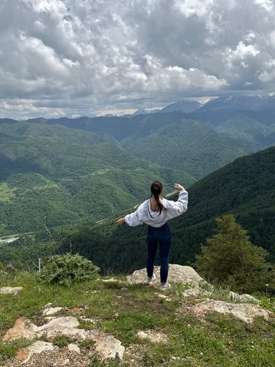 A young woman stands on the edge of a rocky cliff overlooking a vast, green valley filled with rolling mountains. The woman is seen from behind, wearing a grey hoodie and blue jeans, with her arms outstretched as if embracing the expansive landscape. The sky above is partly cloudy, with dramatic grey clouds interspersed with patches of blue, hinting at a dynamic weather system. In the distance, snow-capped mountains form a majestic backdrop. The foreground is characterized by rugged terrain with scattered rocks and green grass. The image captures a moment of solitude and awe in a breathtaking natural setting, potentially in the vicinity of Nizhnyaya Teberda, Russia, a region known for its stunning mountain scenery.