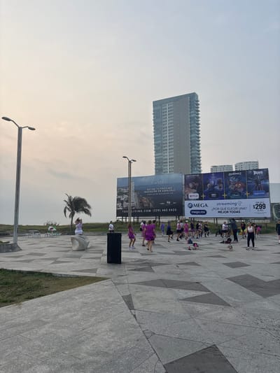 This media file captures an outdoor fitness class or group exercise event taking place on a patterned tiled plaza in Veracruz, Mexico, likely during late afternoon or early evening under a hazy sky.
In the foreground, a group of approximately fifteen to twenty people, mostly women dressed in activewear (some in purple attire), are engaged in a synchronized exercise activity, resembling a dance or aerobics class. They are spread across the patterned grey and dark grey tiled ground. A black rubbish bin is visible near them. Further back, a solitary palm tree stands beside a modern, sculptural white bench.
The background features a modern urban landscape with several tall, multi-story buildings. Dominating the middle ground are two large billboards. The left billboard advertises real estate: "TAURI," "DEFINE TU PRÓXIMA INVERSIÓN EN VERACRUZ," and "COSTA DE ORO, BOCA DEL RÍO, VER.," along with "TAU LIVING.COM (229) 465 0425." The right billboard promotes a streaming service called "MEGA," offering various platforms like Netflix (with "BRIDGERTON"), HBO Max (with a Champions League logo), Amazon Prime (with "BOYS"), Disney+ (with "ZOOTOPIA 2"), and asking "¿POR QUÉ ELEGIR UNA? MEJOR TODAS" (WHY CHOOSE ONE? ALL THE BEST), for "POR SÓLO $299 AL MES" (FOR ONLY $299 PER MONTH). Two streetlights are also visible, one in the lower left foreground and another further back on the left side, adding to the urban setting. Sections of green grass line the edges of the tiled plaza.