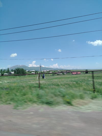 An exterior, ground-level, full shot captures a hazy, daytime scene of a rural landscape with a village and a mountain in the distance. Three black power lines stretch across the clear blue sky. The foreground features a blurred, grassy field with scattered yellow wildflowers and a dirt path or road running horizontally. A rusty brown metal fence runs along the middle ground, separating the field from the village. The village consists of several buildings with red and white roofs, interspersed with green trees. A tall utility pole with a gray electrical box is visible to the right of center. In the background, a large, light-colored mountain with patches of snow rises against the horizon. The overall impression is one of a serene, pastoral setting in the city of Hartavan, Armenia.