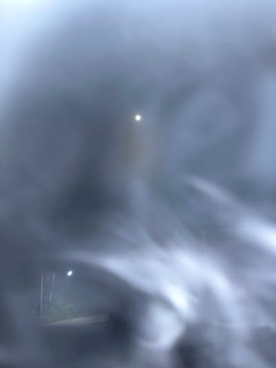 The image depicts a foggy or misty environment with low visibility. In the upper portion, a bright, circular light source, possibly the moon or a strong artificial light, is visible through the haze. The lower part of the image shows faint outlines of what appear to be streetlights and possibly some foliage, suggesting an outdoor setting at night or in low light conditions. The overall atmosphere is obscured by a pervasive fog or mist, making it difficult to discern specific details of the environment. The location in Legonipah, Indonesia, is not visually discernible from the provided image due to the lack of specific landmarks or identifying features. The image appears to have been taken through a wet or smudged surface, contributing to the blurred and indistinct quality of the scene.