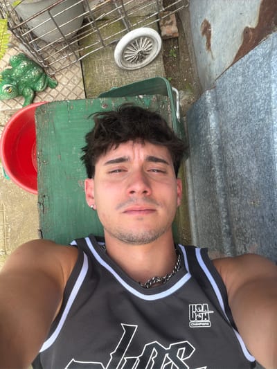 An overhead selfie captures a young adult male lying outdoors in Krumovo, Bulgaria. The man, appearing to be in his 20s, has dark, disheveled hair, a light mustache, and stubble, with a silver hoop earring in his left ear. He wears a dark grey sleeveless basketball-style jersey with white and light grey stripes along the collar and armholes, a silver chain necklace, and a white "USA CHAMPIONS" logo. His gaze is directed at the camera with a neutral expression. He is positioned against a weathered green wooden surface. To the left, on a concrete and tiled ground, are a partially visible red plastic bucket, a green frog statue, a metal cage-like object, and a white spoked wheel. To the right, a corrugated metal sheet with rust stains stands vertically, bordered by dirt ground with sparse vegetation. The scene is brightly lit by natural light.