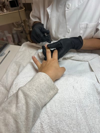The image depicts an indoor scene in Lordelo do Ouro e Massarelos, Portugal, focusing on a manicure service. A person's right forearm, clad in a light grey long-sleeved garment, and their hand rest on a white terrycloth towel that covers a table surface. A second individual, wearing a white uniform jacket with two visible black buttons and black nitrile gloves, is performing a nail treatment. The gloved hands of the second person are actively holding the client's index and middle fingers. A silver-colored nail tool, consistent with cuticle nippers, is visible in the manicurist's left gloved hand. The background is softly blurred, revealing shelves or a trolley with multiple small bottles and containers, indicative of a professional nail salon setting.