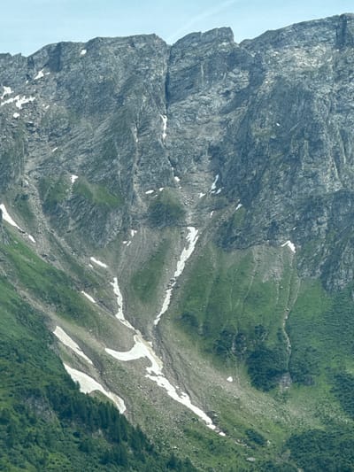 The image depicts a steep, rocky mountain face with patches of green grass and snow. The mountain appears to be covered in scree and has a rugged, uneven texture. Some areas show evidence of erosion or geological formations. The sky above is a light blue. The image does not contain any people, objects, or actions. The scene is a natural mountain landscape.
