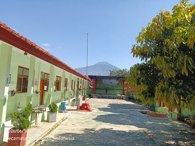 The image shows an outdoor scene at a school in Kuningan, Indonesia. A light green building with a red tile roof runs along the left side of the frame, featuring several windows and doors. In the foreground, there are potted plants and a paved walkway. A large tree with green and yellow leaves occupies the right side of the image. In the background, a stage with a painted backdrop is visible, and further in the distance, a mountain rises against a clear blue sky. The date stamp "2026/06/30 09:57" and location "Kecamatan Kuningan, Indonesia" are overlaid on the bottom left of the image. There are no people visible in the scene.