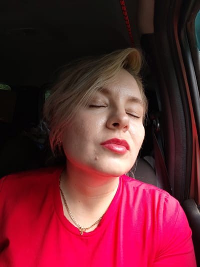 A close-up, eye-level, medium shot captures a fair-skinned woman with blonde hair, her eyes closed and lips pursed, in what appears to be the interior of a vehicle. She is wearing a bright red t-shirt and a gold chain necklace with a small pendant. Her skin shows some texture and a single mole is visible on her left cheek. The background is dark and out of focus, with hints of car interior elements like a seatbelt and window frame. There is a red cord visible overhead. The lighting suggests it is daytime, possibly with sunlight filtering in from the side. The woman's posture and facial expression convey a sense of relaxation or enjoyment. The location is not identifiable from the image itself.