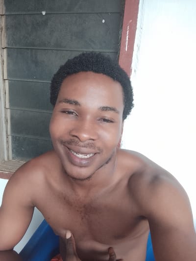 This is a chest-up selfie of a young Black man with short, curly hair. He is smiling, showing his teeth, and looking directly at the camera. He is shirtless, and his skin tone is brown. His left shoulder and upper chest are visible. He has a light mustache and goatee.

The background is partially visible. To the left, there is a window with horizontal blinds, appearing to be made of dark wood or metal. To the right, there is a white wall with a reddish-brown trim along the top, suggesting an outdoor or semi-outdoor setting. The lighting suggests it is daytime. The man appears to be in a relaxed and happy mood.

The provided location context is Orodo, Nigeria.