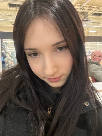 An image taken in a commercial establishment in Mundolsheim, France, features a young woman in the foreground and an older man in the background. The primary subject, a young woman with long, dark brown hair, occupies the majority of the frame, facing slightly downwards and to her left. Her eyes are dark brown and directed downwards. She is wearing a dark, textured garment, possibly a jacket, with a gold-colored metallic accessory visible near her collar. In the mid-ground, to the right of the frame, an older bald male wearing eyeglasses and a light-colored jacket is seated at a table, with a red chair back visible behind him. The background reveals an indoor commercial environment with a high ceiling featuring exposed white structural elements, utility conduits, and fluorescent lighting fixtures. Partially visible on the far left are retail shelving units with various products.