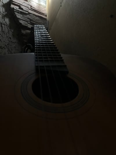 The image shows a low-angle, close-up view of an acoustic guitar. The guitar's neck and fretboard are prominently displayed, stretching upwards towards the top of the frame. The soundhole, with its circular inlay, is visible in the lower center of the image. The body of the guitar is made of wood with a warm, brown hue. The background consists of textured stone walls on the left, illuminated by a light source from above, and a wooden surface on the right, which appears to be a wall or paneling. The overall lighting is dim, creating a moody and atmospheric scene. The image appears to be taken indoors, in a confined space. There is no indication of the location being in Slov'yanoserbs'k, Ukraine, or any other specific city.