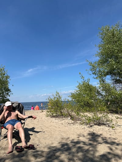 In the city of Demydiv, Ukraine, a man sits in a foldable chair on a sandy beach. He is wearing a white cap and blue shorts. His left arm is resting on the armrest of the chair, and his right hand is near his face. Flip-flops are on the sand in front of him. In the background, the water of a lake or river is visible, with several people in the distance. One person is on a paddleboard, and two others are sitting near the water's edge. Trees and bushes line the sandy area, casting shadows on the ground. The sky is clear blue with a few wispy clouds.