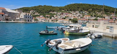 The image depicts a harbor scene in the town of Pučišća, Croatia, on a sunny day. The foreground features several boats moored in clear, turquoise water. Two white motorboats with outboard motors are prominently positioned, with one partially obscuring the view of another. A dark green jet ski is also visible in the water to the left of the motorboats. In the middle ground, a yellow and white boat with a cabin is docked alongside a stone quay. Other smaller boats are also moored in the harbor. The background reveals a hillside densely covered with green trees, dotted with numerous houses with terracotta roofs, characteristic of Mediterranean architecture. The stone buildings along the waterfront in the left of the frame, some with balconies, suggest a historic settlement. A stone monument and electrical boxes are present on the quay. The sky is a clear, bright blue with a few wispy clouds. The overall impression is one of a peaceful, coastal village on a warm summer day.