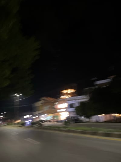 This image depicts a blurred nighttime scene, likely in an urban or semi-urban setting. The composition is dominated by dark areas, suggesting the absence of direct light, with streaks of light indicating artificial illumination and motion.

On the right side of the frame, a road surface is visible, with faint lines suggesting lane markings. Adjacent to the road, there appears to be a grassy or vegetated area, with a yellowish line running parallel to the road, possibly a curb or boundary. Further to the left, building structures are suggested by indistinct shapes and illuminated windows or signs, exhibiting a warm, golden glow. These lights are elongated and streaked, indicating a long exposure or movement during the capture.

Dark foliage, possibly trees or bushes, occupies a significant portion of the left and bottom areas of the image, further contributing to the nighttime ambiance. The overall impression is one of a street scene at night, possibly with cars or people moving, as evidenced by the light trails. The blurred nature of the image prevents the identification of specific individuals or objects, but the presence of illuminated buildings and a road strongly suggests a populated area. The time of day is clearly night. There are no discernible text elements in the image. The scene's mood is difficult to ascertain due to the blurriness, but the artificial lights suggest activity and life despite the darkness. Given the location context of Dalam, Indonesia, this could be a street in a town or city within that region.