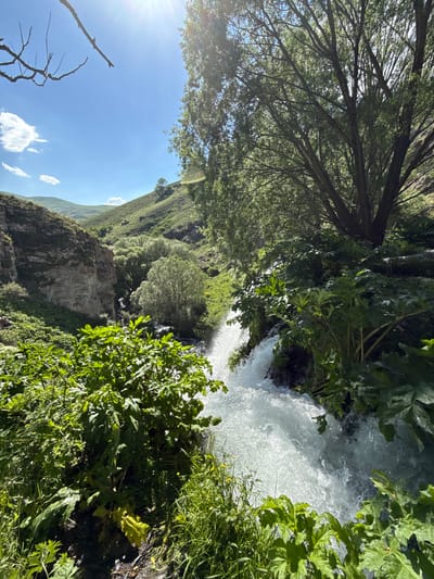 The image captures a natural landscape in the vicinity of Sisian, Armenia. A waterfall cascades down rocks into a pool, surrounded by lush green vegetation and trees. The sun shines brightly through the branches of a large tree in the foreground, casting dappled light on the scene. The background shows rolling hills under a clear blue sky with scattered clouds. Two small figures can be seen standing near the waterfall, observing the natural beauty.