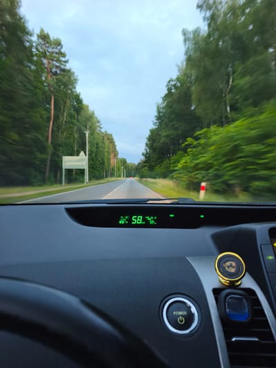 This is a first-person perspective photograph taken from inside a vehicle driving on a road lined with trees. The time of day appears to be dusk or early evening, with a cloudy sky. The road is paved and has lane markings. A digital display on the car's dashboard shows the speed as 58 km/h and indicates the vehicle is in "READY" mode. The interior of the car is visible in the foreground, including a portion of the steering wheel, the dashboard, and a "POWER" button. A decorative magnetic phone holder with a crest design is attached to the center console. The environment outside the car consists of a dense forest on both sides of the road, with tall trees and undergrowth. In the distance, there is a red light visible on the road, suggesting another vehicle. A white signpost with a blank rectangular panel is visible on the left side of the road. A red and white striped marker post is also visible on the right side of the road. The location is identified as the city of Pilchowo, Poland. There are no people visible in the image, and the action is the vehicle in motion.