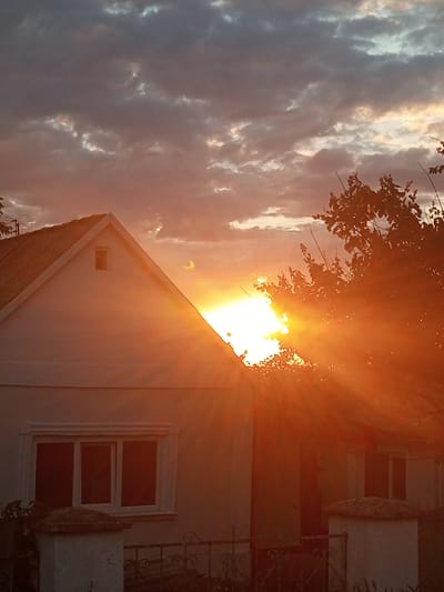 The image depicts a sunset over a residential building in Ароматновское сельское поселение, Ukraine. A white house with a gabled roof and a small attic window is visible on the left. A tree with dark leaves partially obstructs the view of the right side of the house, which also has windows. The sun is setting behind the tree, casting strong orange rays that illuminate the house and clouds. The sky is filled with a mixture of dark and light clouds, with the setting sun creating a bright, warm glow. In the foreground, parts of a fence and two white pillars are visible. The scene is captured in a slightly low-angle perspective, emphasizing the sky and the warm light of the sunset.