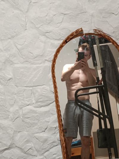 A young man, with light brown hair and a light complexion, is reflected in a cracked, oval mirror. He is shirtless and wearing grey athletic shorts, and he is holding a black smartphone up to take a selfie. His physique is muscular, with defined abs and pectorals. The background of the reflection shows a dark, industrial-looking structure, possibly gym equipment, and some clothing hanging. The mirror has a woven, light brown frame and is positioned in front of a textured white wall that appears to be a rough plaster or stone finish. The setting is likely a personal space such as a gym or home. The image does not contain any information that would help to identify its location in the city of Sucha Beskidzka, Poland.