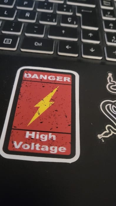 The image displays a close-up, angled view of a section of a dark-colored laptop. A rectangular sticker, featuring a red background with black distressed textures and a white border, is affixed to the laptop's palm rest area. The sticker prominently displays the word "DANGER" in white uppercase letters at the top, a yellow lightning bolt symbol in the center, and the phrase "High Voltage" in white uppercase letters at the bottom. Above the sticker, a portion of the laptop's black keyboard is visible, showing keys with white legends, including "Ctrl" and various arrow keys. To the right of the red sticker, a partial white outline of a second sticker, consistent with the Razer brand logo, is present on the laptop surface. No people are depicted. The scene occurs indoors under ambient lighting. The image provides no visual information to determine its specific location within the city of Magaluf, Spain, or any other geographical area.