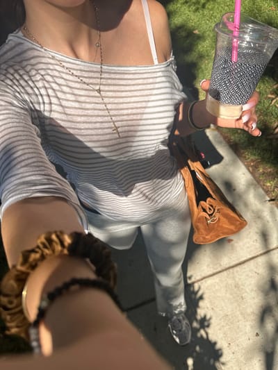 The image depicts a first-person, downward-angled view of an individual walking outdoors in Glendale, United States. The subject wears a sheer, light gray and white striped long-sleeved, off-the-shoulder top, light gray pants or leggings, and gray athletic shoes. A white bra strap is visible on the right shoulder. Two necklaces are worn: a long, gold-toned rosary-style chain with a cross pendant and a shorter chain with a small, circular pendant. The individual's right hand holds a clear plastic cup containing a light-colored beverage, equipped with a pink straw and a black-and-white patterned sleeve featuring the word "COMMISSARY." The fingernails on the right hand are long, pointed, and have a white-to-pink ombré finish. Multiple thin, gold-toned bangles adorn the right wrist. A brown suede-like handbag with a drawstring closure and a gold-toned ring detail hangs near the left hip. The left forearm, prominently in the foreground, displays a brown satin-like scrunchie and a dark beaded bracelet. The environment consists of a concrete or paved sidewalk under bright sunlight, casting distinct shadows from overhead foliage onto the ground. Green grass is visible to the right of the path.