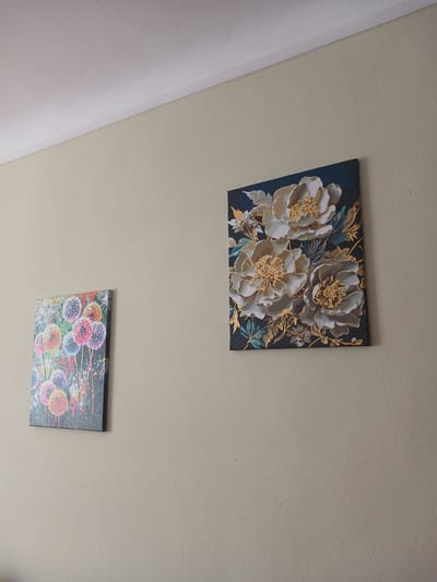 Two framed artworks hang on a light beige wall. The artwork on the left is a vertical canvas depicting colorful, abstract dandelion-like seed heads. The artwork on the right is a square canvas featuring a close-up of three large, stylized white and gold flowers against a dark blue background. The wall is capped by a white crown molding and a white ceiling is visible at the top of the frame. The image appears to be an interior shot, possibly a home or an office, located in Mamaivtsi, Ukraine.