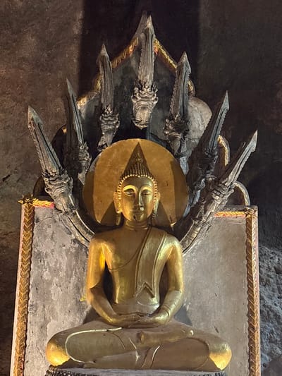 The image displays a golden Buddha statue centrally positioned within a dimly lit cave or rock temple, located in Ban Hin Sam Kon, Thailand. The Buddha is depicted in a seated meditative posture (Dhyana Mudra), with legs crossed and hands resting in the lap, palms upward. The statue has a radiant gold finish, featuring a serene expression, an ushnisha on the crown of the head, and elongated earlobes. A circular golden aureole with a pointed top frames the Buddha's head. Behind the Buddha, a large, dark grey stone or concrete relief forms a multi-headed Naga (cobra) canopy, with at least seven distinct cobra heads fanning out. This Naga structure exhibits detailed carvings of scales and heads, with subtle gold accents along its base and edges. The surrounding environment consists of uneven, dark rock walls, indicating a natural cave or a structure carved directly into rock. The Buddha statue is illuminated by direct light, contrasting with the darker background.