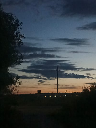 The image shows a dusk sky over a rural landscape in Shestunikha, Russia. Dark silhouetted trees frame the left side of the image, while the horizon is dotted with dark, indistinct vegetation. A tall, slender communication tower stands prominently in the middle ground. In the distance, a low-lying building with illuminated windows is visible, suggesting human habitation or activity. The sky is a gradient of deep blue at the top, transitioning to lighter blues and oranges near the horizon, with scattered dark clouds. The overall impression is one of a serene, twilight scene in a natural setting.