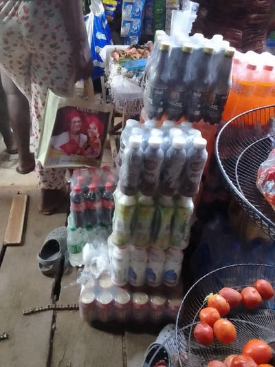 The image shows a street vendor's stall. Stacks of bottled drinks like Coke, Sprite, and chocolate milk tower beside orange soda. A metal basket holds tomatoes, while another contains various root vegetables. A woman in cherry-print pants stands by, carrying a tote bag with a portrait. A grey sandal sits next to a wooden plank, adding to the clutter of the outdoor marketplace.