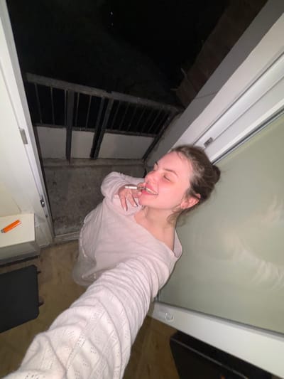 A woman is smoking a cigarette in an alcove. She is fair-skinned with brown hair pulled back in a bun, wearing a light-colored long-sleeved shirt. She has a ring on her right index finger. The alcove has a white-framed window or door, a metal grate, and a lighter on the sill. Beyond the grate, it is dark. The floor is brown and there is a dark-colored object near her feet. The image does not provide any indication that the location is Chornomorskoe, Ukraine.