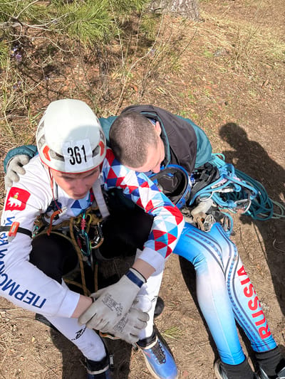 The image depicts two individuals lying on the ground outdoors in Lesnoye, Russia. One individual is wearing a white helmet with the number "361" on it and a white top with a red and white emblem on the sleeve, along with white and red diamond-patterned pants. The other individual is wearing blue and white leggings with the word "RUSSIA" in red letters. Climbing ropes and carabiners are visible around them. The ground appears to be covered with dirt, dry grass, and pine needles.