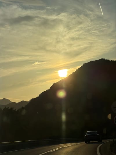 The image shows a landscape during sunset, taken from the city of Gospoština, Montenegro. The sun is setting behind a mountain range, casting a golden light onto the sky and the clouds. A contrail from an airplane is visible in the sky. On the right side of the image, a portion of a road with a street light is visible. The image appears to be taken from within a vehicle or building, as part of the frame is visible in the foreground.