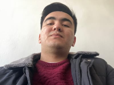 An adult male, estimated to be of Central Asian descent, is depicted in a close-up, chest-up portrait. He possesses dark hair, styled upwards, and a neutral facial expression. Visible on his cheeks are skin textures consistent with acne scarring. He is wearing a dark-colored, possibly navy or black, winter jacket featuring a grey faux-fur or fleece collar. Beneath the jacket, a maroon cable-knit sweater and a white undershirt are discernible. The subject is looking directly into the camera. The background consists of a plain, light-colored wall with minor surface imperfections, indicative of an indoor setting. The scene is located in Namangan City, Uzbekistan.
