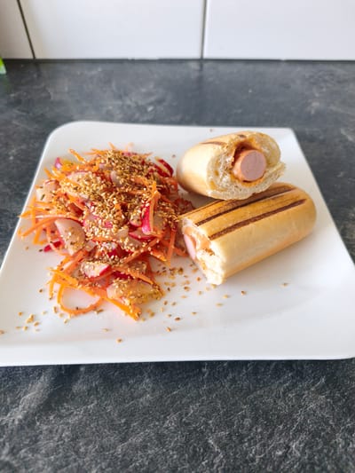 A white plate holds a halved hot dog in a grilled bun and a vibrant salad. The hot dog is split lengthwise, revealing a pink sausage nestled in a creamy orange sauce, with grill marks visible on the bun. The salad is a colorful mix of shredded carrots and thinly sliced radishes, generously sprinkled with sesame seeds. The plate sits on a dark, speckled countertop, likely in a kitchen. The background shows a white tiled wall and a dark grey speckled surface, possibly another countertop or a backsplash. The lighting suggests daytime, with the scene appearing to be indoors. There are no people visible, and the overall mood is neutral and focused on the food. No text is visible in the image.