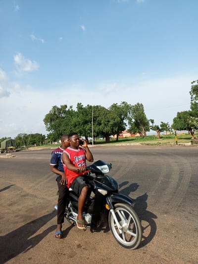 In Gadao, Nigeria, two young men are seated on a black motorcycle. The individual in front, wearing a red Lakers basketball jersey and black shorts, is holding a mobile phone to his right ear, engaged in a conversation. Behind him, another young man, dressed in a blue and white patterned shirt and black pants, is also seated on the motorcycle. The motorcycle is positioned on an asphalt road, casting shadows on the ground. In the background, a truck covered with a green tarp is visible on the left, and lush green trees line the horizon under a bright blue sky with scattered white clouds.