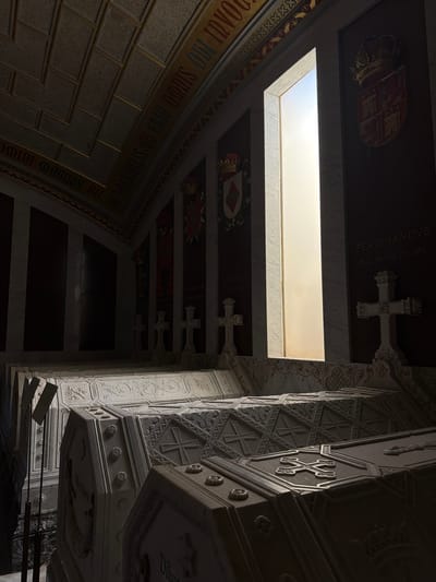 The image depicts the interior of the Royal Pantheon of the Monastery of San Lorenzo de El Escorial, located in San Lorenzo de El Escorial, Spain. The scene features multiple ornate sarcophagi, likely belonging to Spanish royalty, arranged in a dimly lit chamber. The sarcophagi are made of white marble and are elaborately carved with intricate patterns, crosses, and floral motifs. Some of the sarcophagi have inscriptions, though they are largely unreadable in the given lighting conditions. The walls are dark, with some areas featuring marble panels and decorative elements like coats of arms and inscriptions. A prominent feature is a tall, narrow window with light filtering through, illuminating a section of the chamber and casting shadows. Several crosses are positioned on pedestals near the sarcophagi, adding to the solemn atmosphere of the burial site. The ceiling is vaulted and appears to be constructed with a textured material, possibly stone or stucco, with decorative bands. The overall impression is one of grandeur, solemnity, and historical significance, characteristic of a royal mausoleum. There are no people visible in the image.