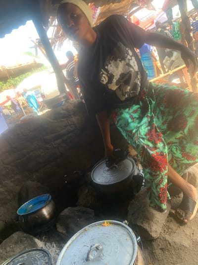 A person wearing a black t-shirt with a white graphic and a light-colored head covering is seen from the chest up, looking towards the camera. They are sitting and their arm is raised, hand extended upwards. Their legs are crossed, and they are wearing a green and red floral patterned skirt or wrap. To the right of the person, a large pot is visible, sitting on what appears to be rocks. There are other pots and metallic objects around. The scene appears to be outdoors, possibly a market or a rural setting, with structures made of wood and what looks like corrugated metal or palm fronds overhead. The lighting suggests it is daytime. In the background, other people are visible, some appearing to be wearing traditional clothing, and there are stalls with various items, including boxes and bags. The ground is sandy and dusty. One of the boxes in the background has visible text which reads "ANTEX M EXCLSY". The overall impression is of a busy, informal gathering or marketplace.