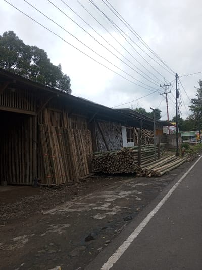 A roadside scene with a weathered, bamboo-built structure displaying stacks of bamboo poles. Some are bundled and leaning against the building, while others are piled in neat stacks. A narrow road runs along the side, and overhead power lines crisscross the cloudy sky. In the distance, hints of greenery and other buildings can be seen. The ground is uneven and shows signs of wear.