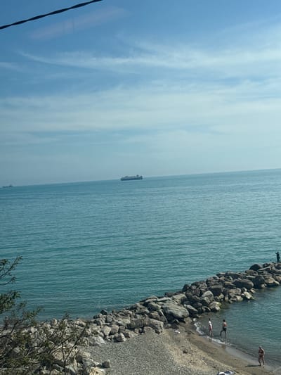 An elevated daytime view captures a coastal scene in Prigorodnyi, Russia, under a clear blue sky with sparse cirrus clouds. A dark, wavy line, identified as either a cable or a reflection, diagonally traverses the uppermost portion of the image. The expansive blue-green sea stretches to the horizon, where two large cargo ships are visible; one is more prominent in the mid-ground, and a smaller vessel is further to the left. In the foreground, a pebbled beach transitions into a prominent rock breakwater extending into the water. Several individuals are present: one person stands on the far end of the rock breakwater with arms raised towards the sea. Near the base of the breakwater, a male and a female in swimwear wade in the shallow water. To the far right, another person in swimwear walks out of the water onto the sand. On the left side of the pebbled beach, partially obscured by green foliage, a lone figure is walking.