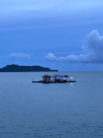 The image depicts a serene scene at dusk in Pa Khlok, Thailand. The primary subjects are a floating structure, possibly a house or raft, on a calm body of water and a silhouetted mountain range in the background. The floating structure is wooden and appears to have a thatched roof on a smaller section, while a larger section has a more modern roof. There are lights visible on the floating structure, suggesting it is inhabited or in use. Small rafts or buoys are also visible near the main structure.
The time of day is clearly dusk, indicated by the deep blue hues of the sky and the soft, diffused lighting. The weather appears to be calm, with the water showing minimal ripples. The sky is partially filled with large, soft clouds, adding to the tranquil atmosphere. The mountain range's dark silhouette against the twilight sky provides a sense of depth and natural beauty to the scene.
The overall mood of the image is peaceful and contemplative. There is no visible text in the image. The scene captures a quiet moment, likely at the end of a day, with the lights on the floating structure offering a hint of human presence in the otherwise natural landscape. The location context of Pa Khlok, Thailand, suggests a tropical or subtropical setting.