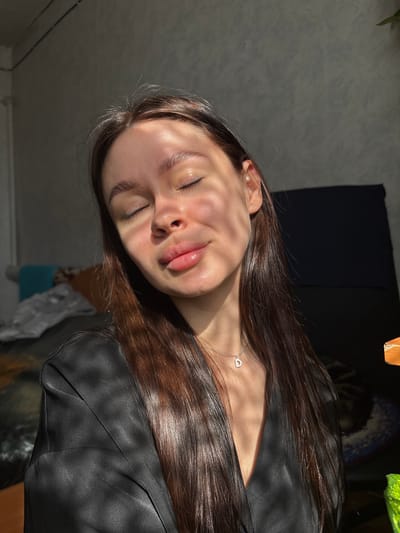 A portrait of a young woman with long brown hair wearing a black shirt, and a silver necklace with a heart-shaped pendant. She has her eyes closed and is facing upwards. Sunlight casts shadows across her face. The background includes a dark wall, a plant, and a glimpse of a room with a wall and possibly a piece of furniture. No information regarding the location in the city of Dubrova, Russia is available.