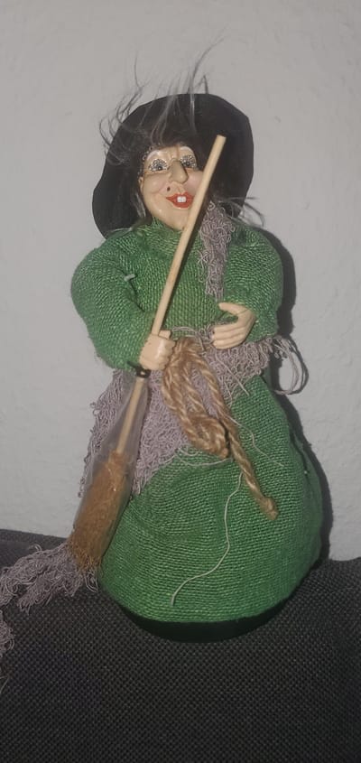An articulated doll, designed to resemble a witch, stands upright against a textured off-white wall. The doll is wearing a textured green dress and a black hat. It has grey hair that appears to be wispy. The doll's face is painted with exaggerated features, including wide eyes with sparkly accents, a prominent nose, and red lips. In its hands, the doll is holding a broomstick with a light brown handle and straw-like bristles enclosed in clear plastic. A textured, light purple scarf is draped around the doll's shoulders, and a thick, twisted rope is tied around the broomstick. The doll is positioned on a dark grey, textured surface. The image does not provide any information or context related to the city of Sasbach, Germany.