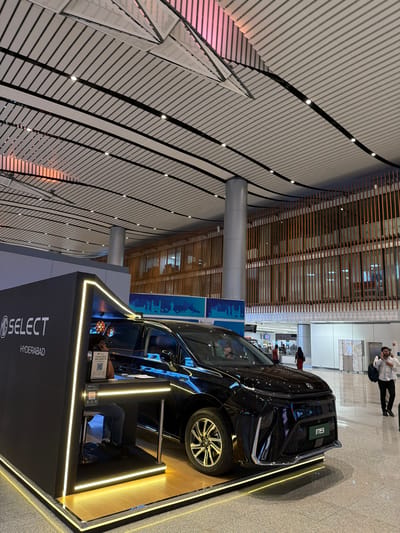 The image shows a black MG electric car on display at what appears to be an airport terminal. The car is positioned in front of a black display booth with "MG SELECT HYDERABAD" written on it, illuminated by neon yellow lights. A person is seated inside the booth, possibly an employee attending to visitors, with a QR code visible on a stand. The ceiling of the terminal has a distinctive modern design with angled slats and integrated lighting. In the background, a multi-story building with wooden vertical slats is visible, and several people are walking through the terminal. The floor is polished and reflects the surrounding lights and structures. The location is identified as Gollapalle Khurd, India.