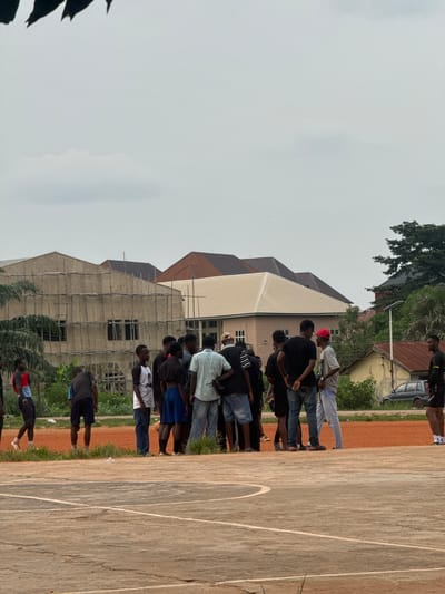 In Amansee, Nigeria, a group of men are gathered on an outdoor court, possibly a basketball court given the markings. The court appears to be made of concrete and is surrounded by a reddish dirt area with some patches of grass. In the background, several buildings are visible. One prominent building is under construction, with scaffolding made of bamboo poles. Other buildings have distinctive tiled roofs in shades of brown and beige. Lush green vegetation, including palm trees and other foliage, surrounds the buildings and the court. The sky is overcast and cloudy. The men are dressed casually in a variety of t-shirts, shorts, and jeans. Some appear to be conversing, while others are standing and observing. One man on the far right is wearing athletic shorts and appears to be in motion or just finished an activity. The overall scene suggests an informal gathering or a break during an outdoor activity.