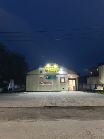 The image captures a modest, light-yellow corrugated metal building at dusk in Vilyuchinsk, Russia. The building appears to be a small shop or establishment, with "ПЕННАЯ БУХТА" (Foamy Bay) prominently displayed in yellow lettering on a banner above the entrance. Below this, smaller text indicates offerings like "ПИВО, РЫБА, ПРОДУКТЫ" (Beer, Fish, Products) and "КОФЕ, НАПИТКИ, ВЫПЕЧКА" (Coffee, Drinks, Baked Goods). A red sign on the wall reads "МЫ ОТКРЫЛИСЬ!" (We Are Open!).

The entrance to the building is illuminated, with a single door ajar, revealing a dimly lit interior and a counter. A small air conditioning unit is mounted above the door. To the left of the building, a white truck with a blue and white cargo box is parked on a gravelly area. In front of the building, a paved walkway made of square grey tiles leads to the entrance.

The environment is residential or commercial, with a dark blue sky suggesting late evening. A few trees are visible to the left, silhouetted against the sky. To the right of the building, a pale yellow fence encloses an area with some greenery, and a wooden bench sits on the paved area. The foreground shows a dark asphalt road. The overall impression is of a small, local business operating in a quiet, possibly suburban, area of Vilyuchinsk.
