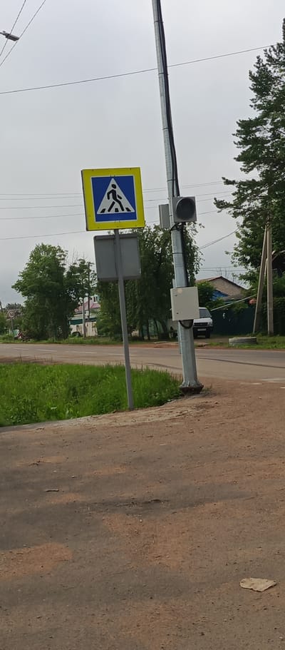 The image shows a pedestrian crossing sign on a pole, a traffic signal, and a speed limit sign in the city of Pogranichny, Russia. The pedestrian crossing sign is yellow and blue with a white silhouette of a person walking. The traffic signal is black and mounted on the same pole as the pedestrian crossing sign. A speed limit sign with the number "50" is visible in the background to the left. The scene appears to be on a cloudy day, with lush green grass and trees surrounding the road. A white van is parked on the side of the road in the background.