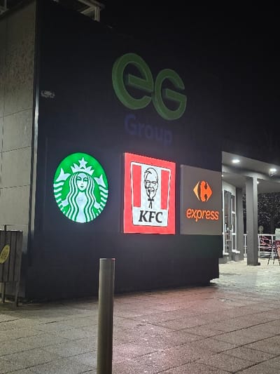 The image displays the illuminated facade of a commercial building at night. Several prominent brand signs are mounted on a dark grey or black wall.
On the left, a large, stylized green logo consisting of two interlocking 'e' letters represents "ee Group," with the word "Group" written vertically in blue alongside it.
To the right of the "ee Group" logo, three additional brand signs are stacked vertically. The topmost sign is rectangular, featuring an orange logo resembling the "Carrefour Express" emblem with the word "express" written in orange lowercase text. Below it is a bright red, square sign for KFC (Kentucky Fried Chicken), displaying the white image of Colonel Sanders and the letters "KFC." The bottommost sign is a circular, green and white illuminated logo for Starbucks, depicting its iconic siren.
The bright illumination of the signs against the dark background confirms it is nighttime. The ground visible at the bottom and to the right is paved with reddish or light-colored bricks or tiles. A metallic pole is partially visible on the right side of the frame. No people or specific activities are depicted in the scene. The overall setting is characteristic of a commercial strip or shopping area, likely a standalone building or part of a larger complex. Given the location context of Longeville-lès-Saint-Avold, France, these are common European and international retail and food service brands.