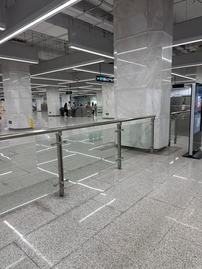 The image depicts an interior scene, likely a modern subway station or public transport hub, in Liwan District, China. The environment is characterized by polished granite flooring with embedded light strips creating a reflective surface. Large, octagonal columns clad in white marble dominate the space, supporting a high ceiling with extensive fluorescent lighting and exposed infrastructure.

In the mid-ground, a transparent glass barrier with stainless steel railings guides pedestrian flow. Beyond this barrier, several people are visible, some interacting with ticket gates or waiting. One group of individuals is gathered near a set of automatic gates, suggesting a point of entry or exit. Another person is walking away from the camera towards the left.

A digital display screen is situated on the right side of the frame, displaying what appears to be promotional content or information. Signage with Chinese characters is visible in the background, including directional arrows and symbols indicating facilities. The overall impression is one of a clean, well-lit, and functional public space.