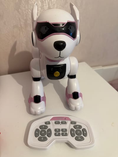 A white robot dog with pink accents and a remote control are displayed on a white surface. The robot dog has a black nose, black and pink digital eyes, and two white ears. A yellow button is visible on its chest. The remote control is white with pink accents and features various grey buttons with icons. The background is a textured, light-colored wall. The scene is not located in Wolfsburg, Germany.