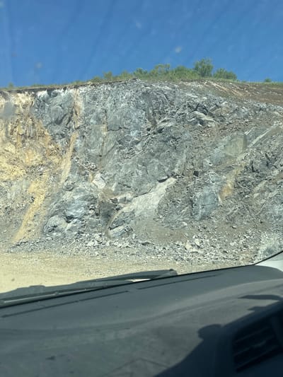 This is a photograph taken from inside a vehicle, looking out at a large rock face and the sky. The rock face is steep and appears to be a quarry or excavation site, with layers of gray and yellowish rock visible. Loose rocks and gravel are scattered at the base of the face. Above the rock face, a strip of greenery with trees and bushes can be seen along the top edge. The sky is a clear, bright blue, suggesting it is daytime and a sunny day. The interior of the vehicle is partially visible in the foreground, with the dashboard and windshield wipers apparent. The image does not depict any people or specific actions. The location is identified as Sarasa, Russia.