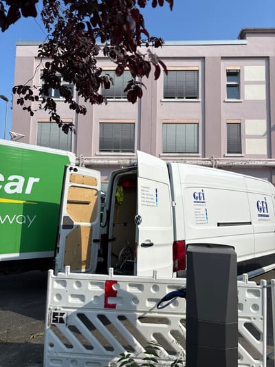 The image shows a street scene in Mannheim, Germany, in front of a pale pink building with blue-tinted windows and horizontal blinds. A large green truck with white lettering that reads "car" and "way" is parked to the left. To the right of the green truck is a white panel van with its rear doors open, revealing the interior with wooden paneling. The van has "GFI GESELLSCHAFT FÜR INDUSTRIETECHNIK mbH & Co. KG" printed on its side, along with contact information including an address "Industriestraße 2, 64653 Lorsch". A yellow safety vest is hanging inside the van. In the foreground, there is a black electric vehicle charging station with a blue charging cable connected to it. A white plastic barrier with red markings is also present in front of the charging station. Dark red leaves from a tree are hanging into the frame from the top left. The sky is clear and blue.