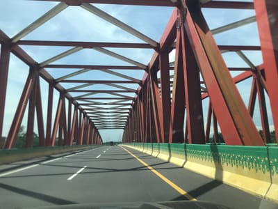 The image shows a view from a car driving across a truss bridge in Gohor Lama, Indonesia. The bridge structure is primarily red, with some silver-colored crossbeams. The road is gray asphalt with white and yellow lane markings. The bridge's guardrail is painted yellow and green. The weather is clear with some clouds visible in the background. Other vehicles are on the bridge. Trees are visible beyond the bridge structure.